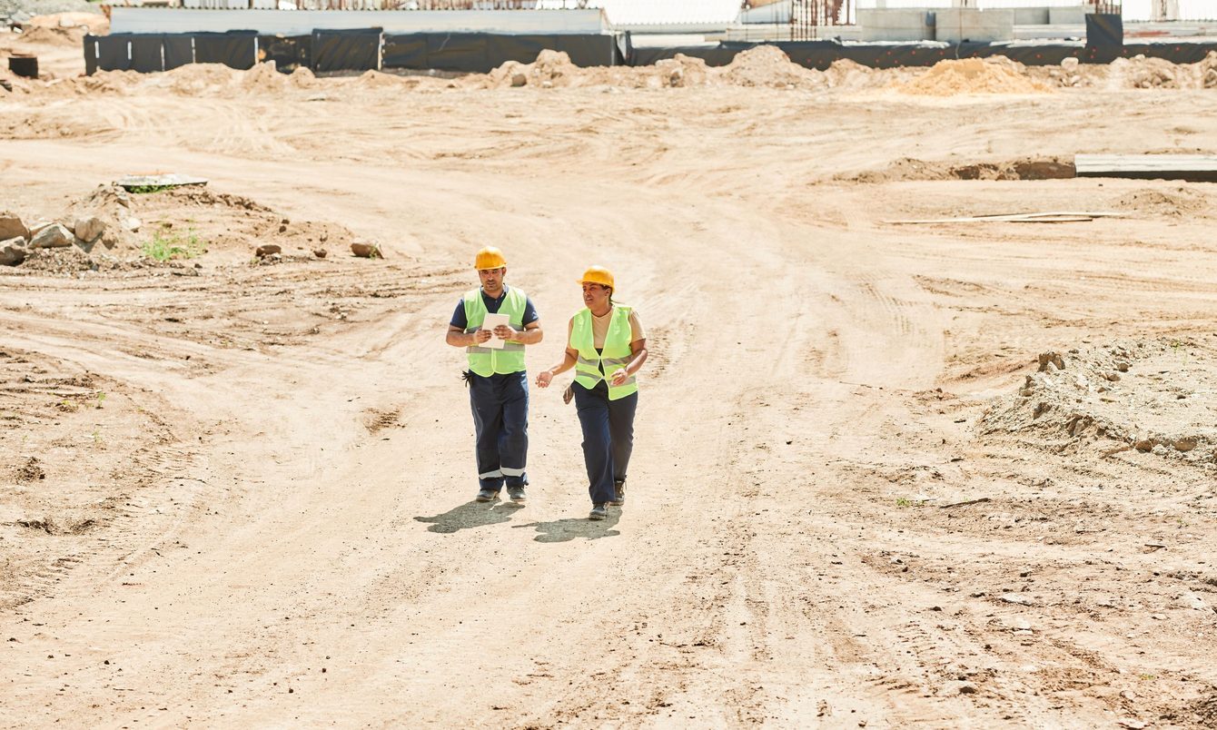 Two people in hard hats and hi-vis vests walking across a graded construction site