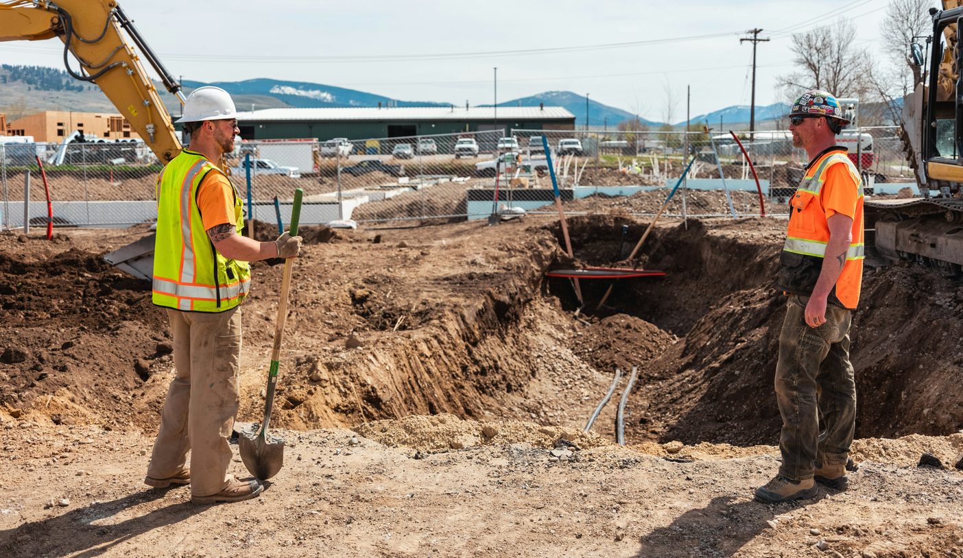 Two workers in hard hats and high-visibility vests standing still on either side of an open excavation on a commercial jobsite, one holding an unused shovel, the excavators behind them stopped