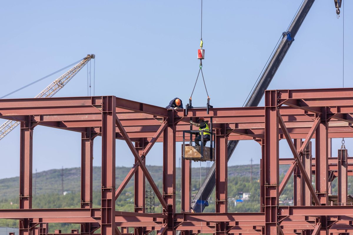 Two ironworkers setting a steel beam from a crane-hung basket on an open frame