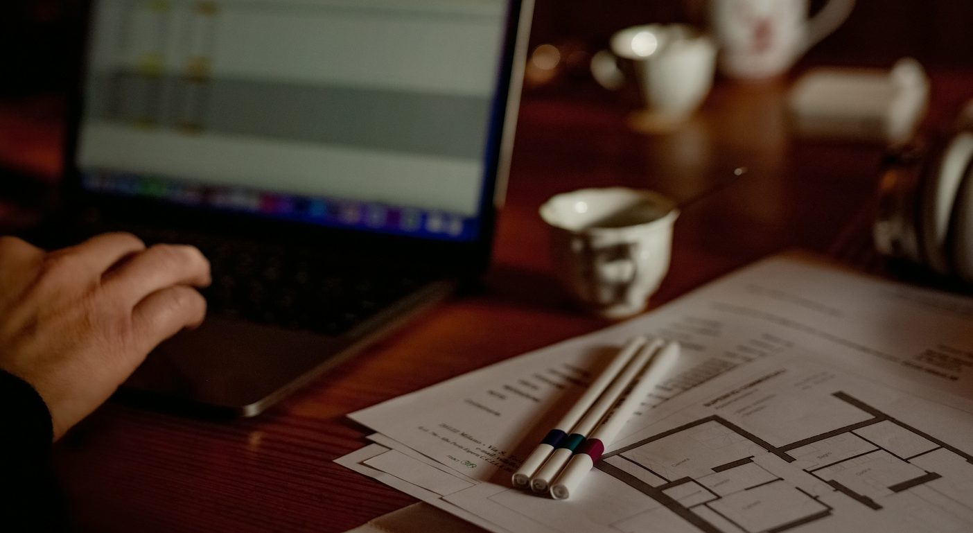 A hand on a laptop trackpad after dark, a spreadsheet open on the screen, beside printed paperwork, a floor plan and marker pens spread across a warm-lit wooden desk